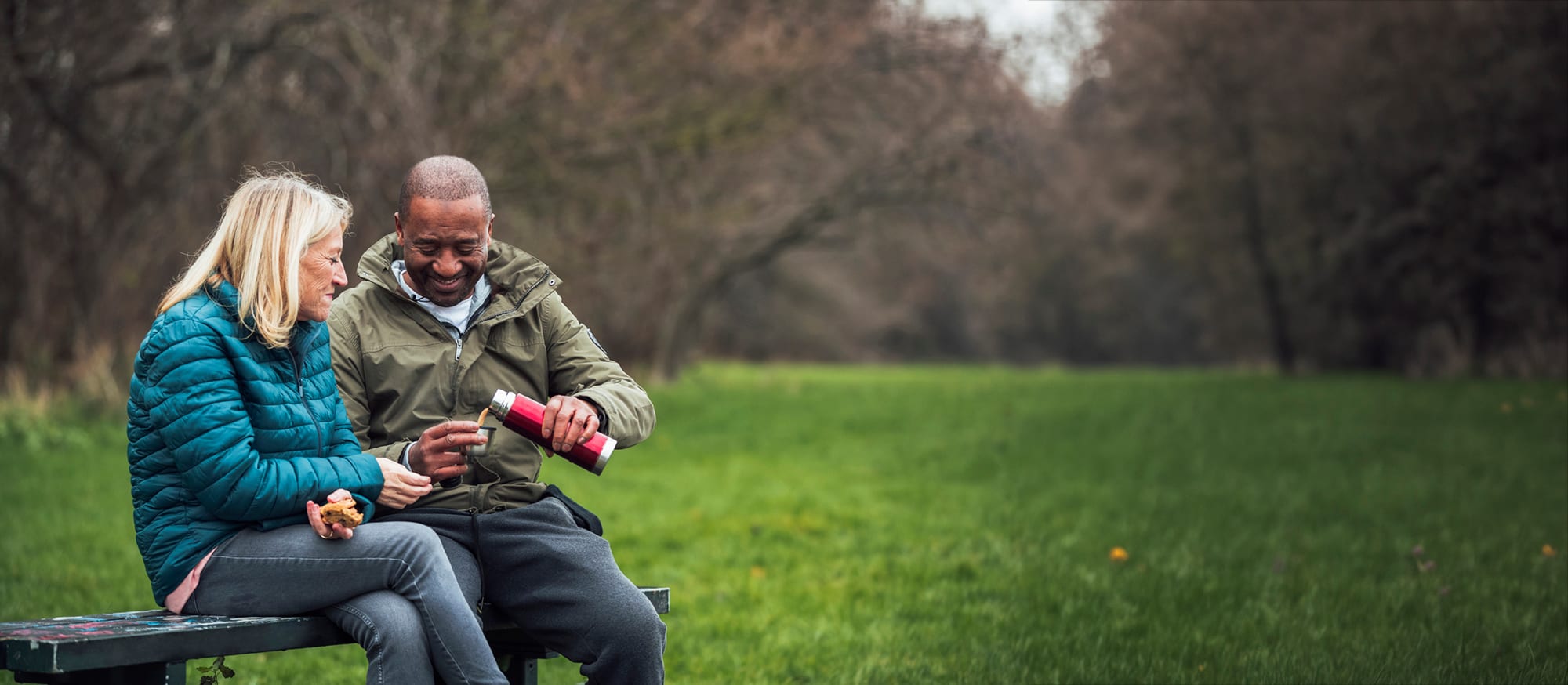 Man and lady in the park share a tea from a flask on the park bench
